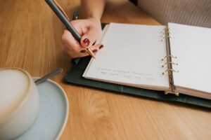 Journaling Fragen in einem Heft. Daneben steht ein Kaffee. Die Frau schreibt Journaling für ihre Persönlichkeitsentwicklung.